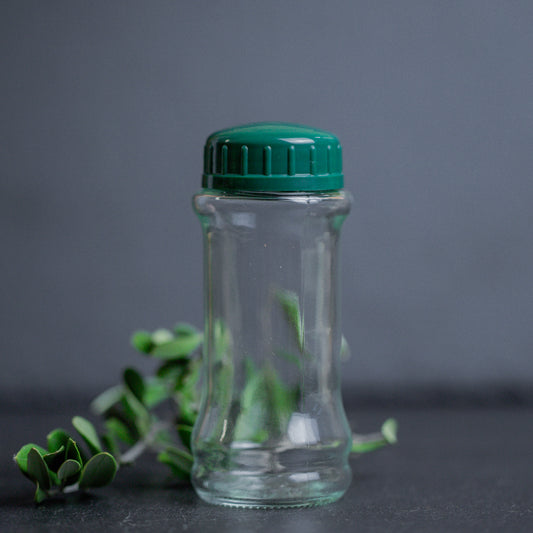 Gewürzstreuer aus Glas mit grünem Deckel auf dunklem Untergrund mit grünblättrigen Zweigen